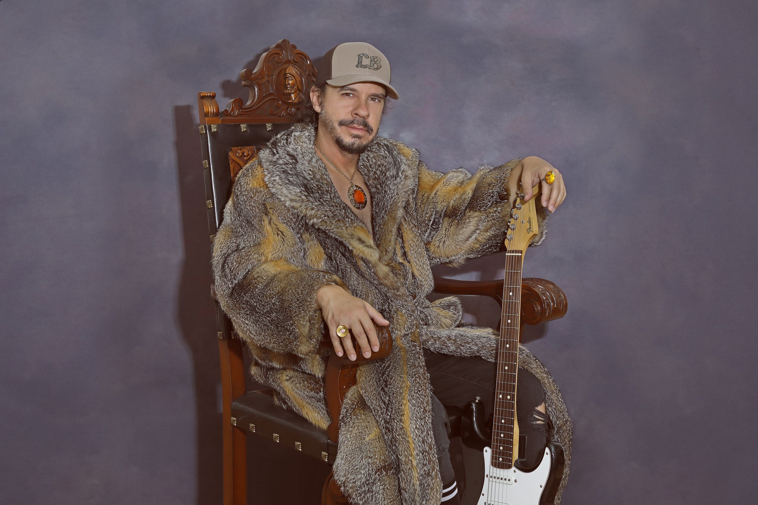 Luis Brea sentado en un trono de madera, con abrigo de piel y una guitarra